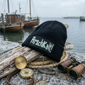 Digital bearbeitete Produktansicht: schwarze Arschkalt!-Mütze auf Treibholz am frostigen Hafen